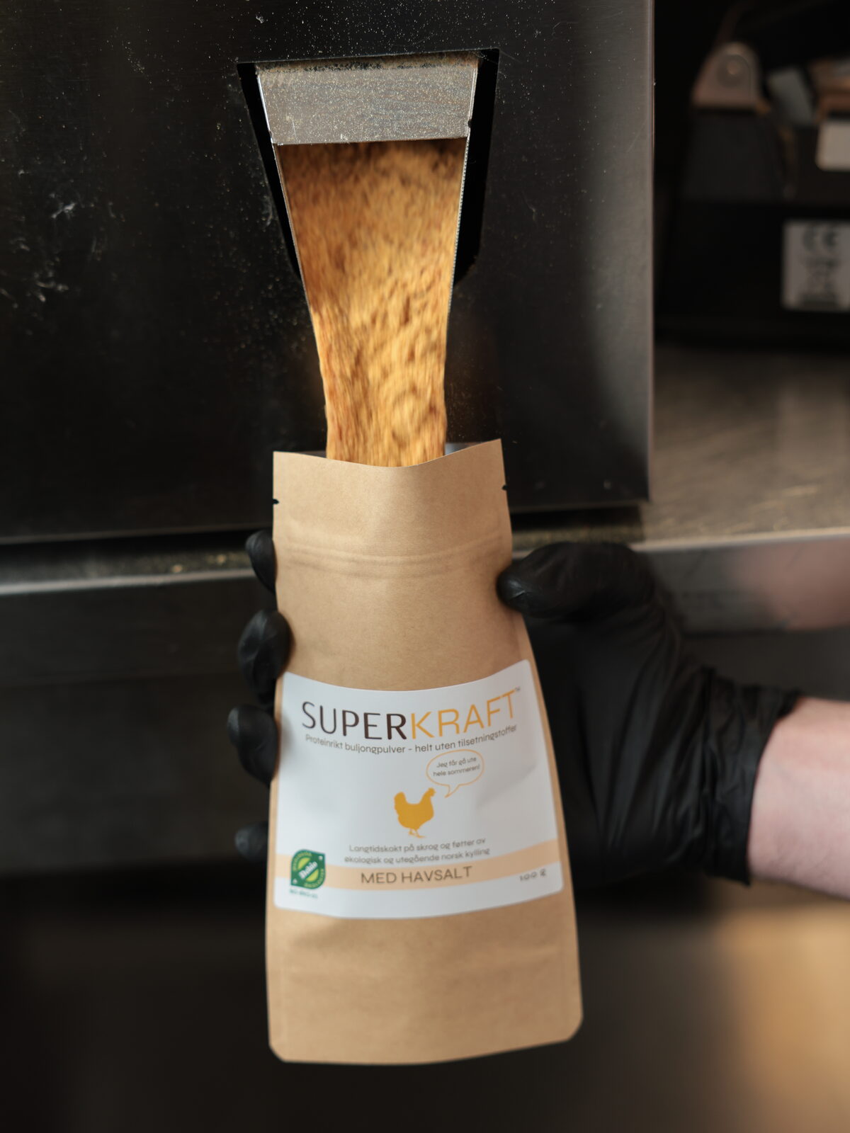 Kraftpulver renner fra fyllemaskinen ned i en pose Superkraft med havsalt, holdt med svarte hansker